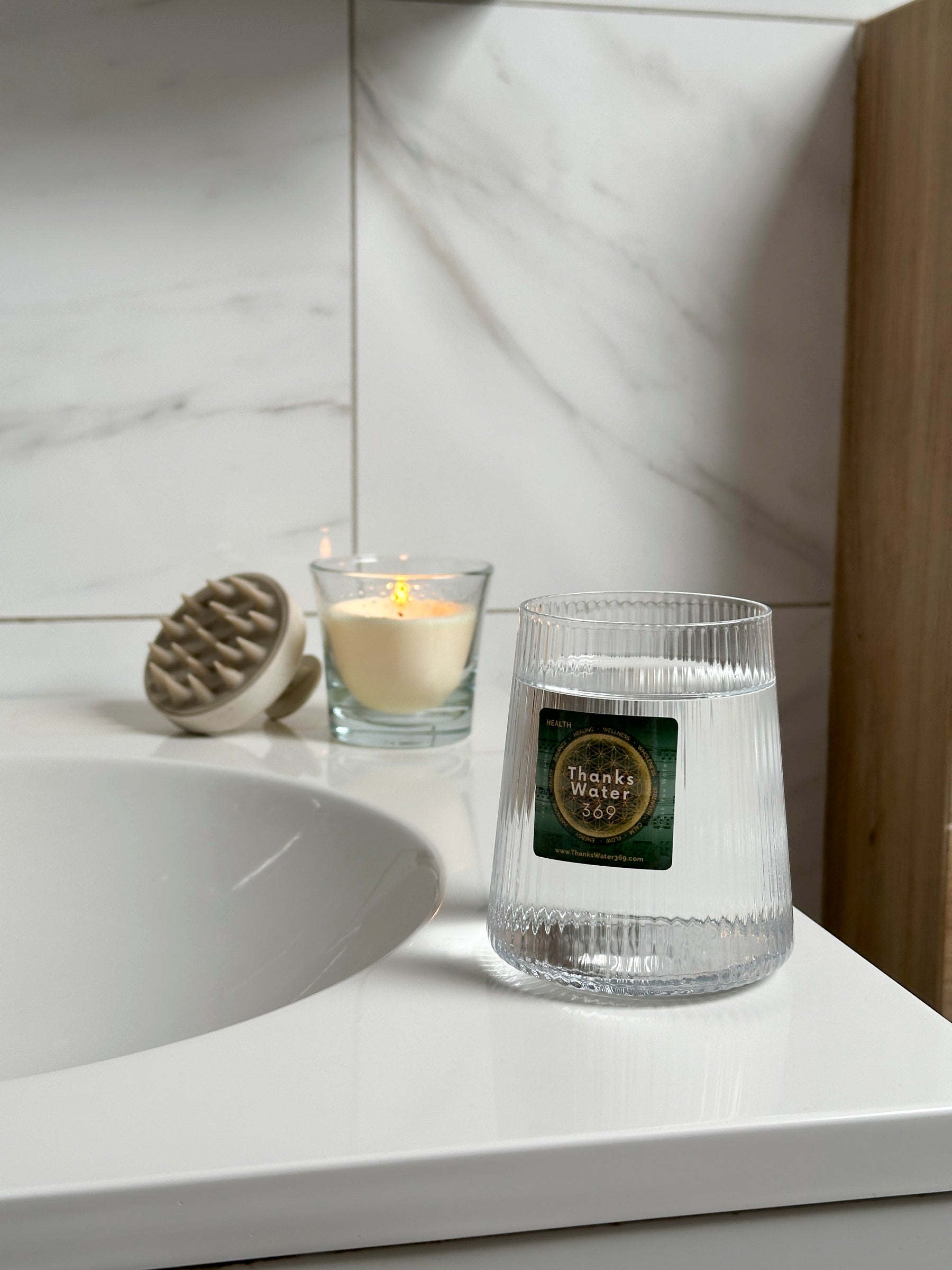 A ThanksWater369 Health label applied to a fluted glass of water on a marble bathroom counter, beside a lit candle