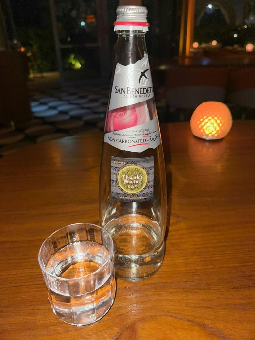 A labelled bottle and glass on a wooden table beside a lamp